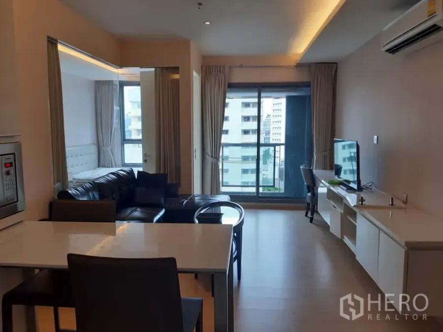 Condominium for rent in Watthana, Bangkok - Bright living room with leather sofa, TV console, and sliding glass doors opening to a city-view balcony.