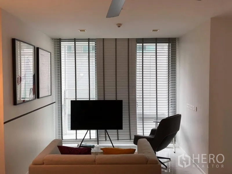 Condominium for rent or sale in Watthana, Bangkok - Living room with sofa, TV on stand, armchair and tall windows fitted with blinds.