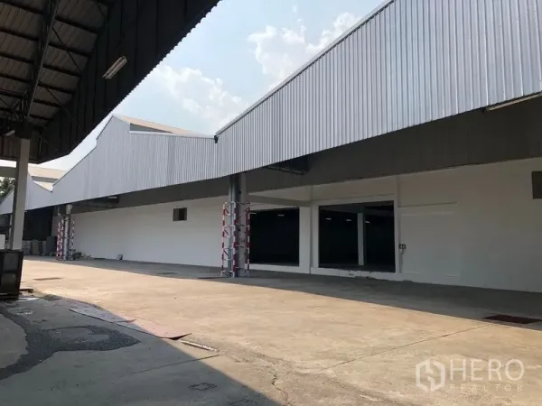 货栈 出租在Pak Kret, Nonthaburi - 仓库外立面为人字形屋顶，带长条雨棚装卸区和宽阔混凝土车道。