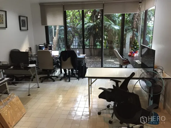 Townhouse for rent in Watthana, Bangkok - Spacious ground-floor room with desks, tiled floor, and full-height windows opening to a leafy garden.