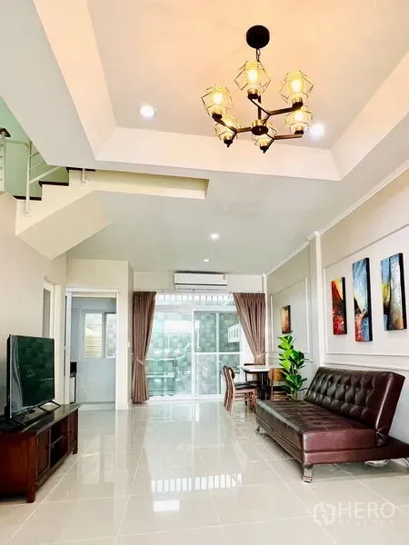ทาวน์เฮาส์ ให้เช่า - ห้องนั่งเล่นโปร่งโล่ง โคมไฟระย้า โซฟาหนังสีน้ำตาล ทีวี บันได และประตูกระจกบานเลื่อน