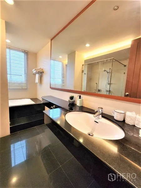 Condominium for rent in Bang Rak, Bangkok - Bathroom featuring a long stone vanity, large mirror, step-up bathtub and window.