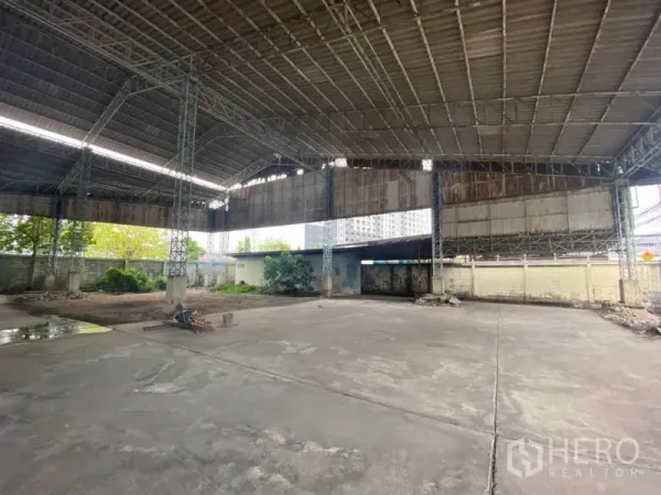 Warehouse for rent in Suan Luang, Bangkok - Wide concrete yard under high steel truss roof with partial walls and greenery.