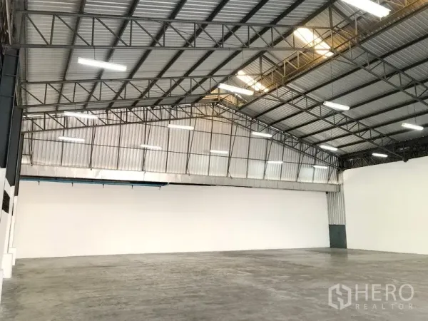 Warehouse for rent in Pak Kret, Nonthaburi - Angled view of open-plan storage hall showing a tall gabled roof, bright lights, and clean concrete flooring.