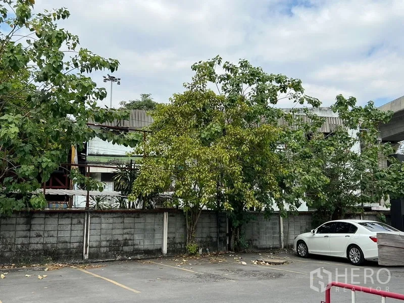 Shophouse for rent or sale in Bang Khen, Bangkok - Parking area beside the property with trees, boundary wall and a parked car.