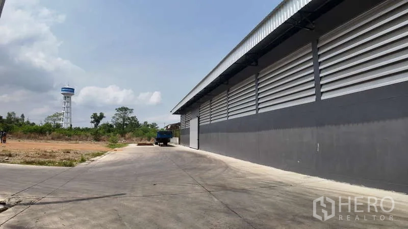 Warehouse for rent in Chachoengsao - Side exterior with concrete apron for trucks and a distant water tower in view.