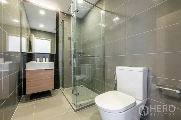 Condominium for rent or sale in Watthana, Bangkok - Modern bathroom with glass-enclosed shower, grey tile walls and floating vanity.