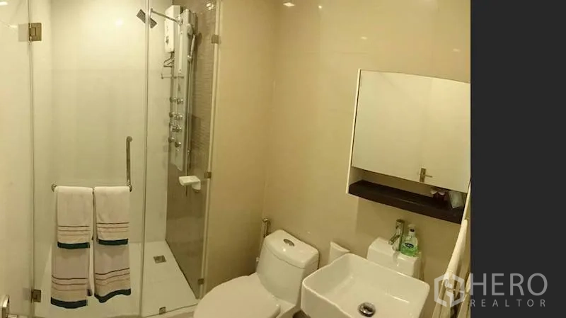 Condominium for rent or sale in Watthana, Bangkok - Compact bathroom with glass-enclosed shower, white vanity and toilet in neutral tiles.