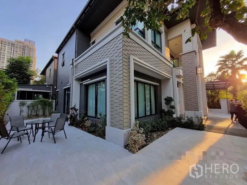 Detached House for rent in Bang Khae, Bangkok - Two-storey house corner with patio seating, garden beds and large windows at sunset.