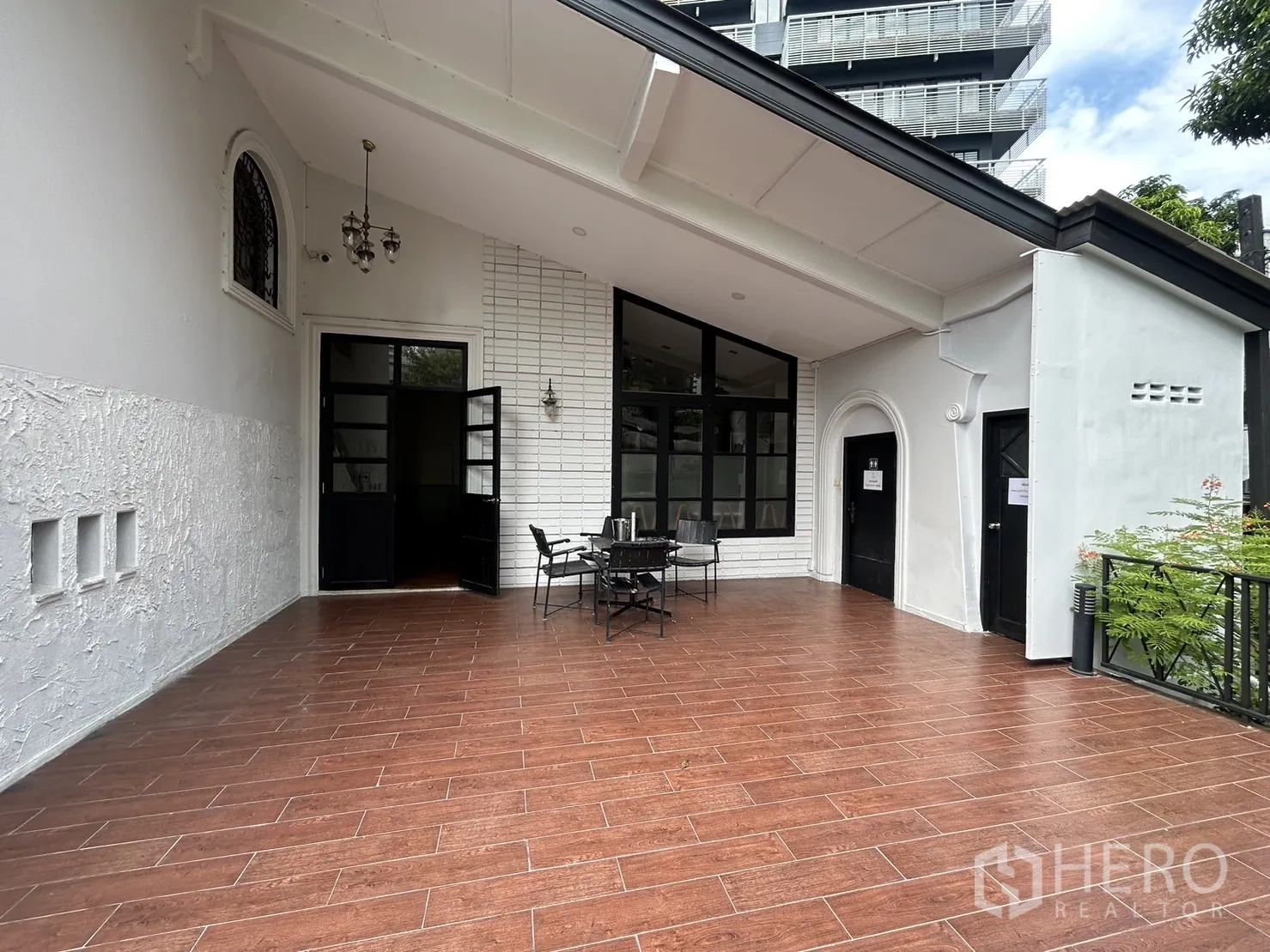 พื้นที่ขายปลีก ให้เช่าในBangkok - ชานระเบียงกว้างปูกระเบื้องลายไม้ ประตูกรอบกระจกสีดำ และชุดโต๊ะเก้าอี้กลางแจ้ง