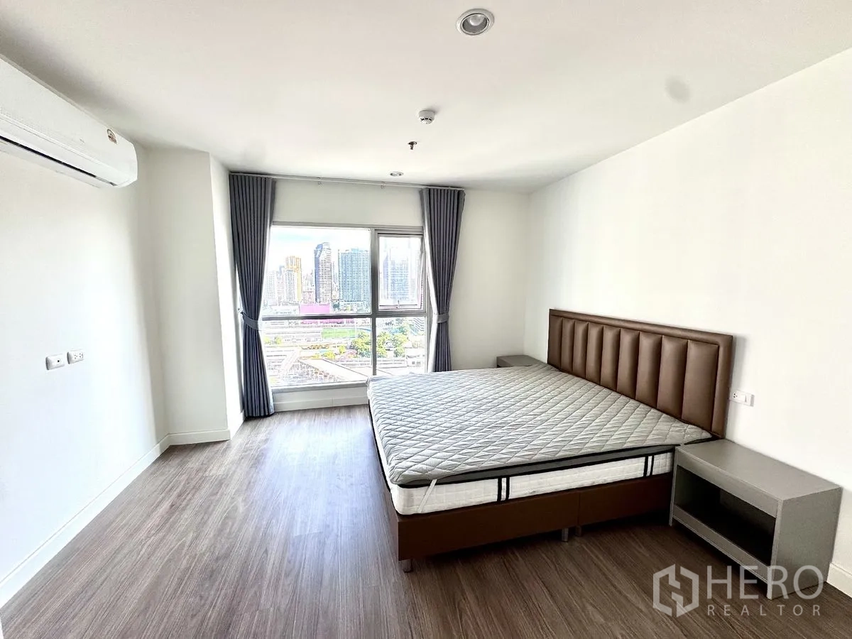 Condominium for rent in Huai Khwang, Bangkok - Spacious bedroom with large window, double bed, bedside tables, wood flooring, and air conditioner.