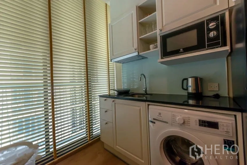 Condominium for rent in Watthana, Bangkok - Compact kitchen with white cabinetry, black countertop, washer and microwave beside tall blinds.