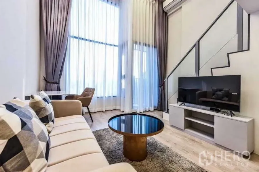 Condominium for rent in Huai Khwang, Bangkok - Sofa seating facing TV under tall curtains and contemporary staircase.