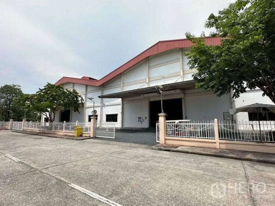 货栈 出租在Bang Sao Thong, Samut Prakan - 白色仓库配红色山墙屋顶，前方有围栏、宽阔车道与装卸雨棚。