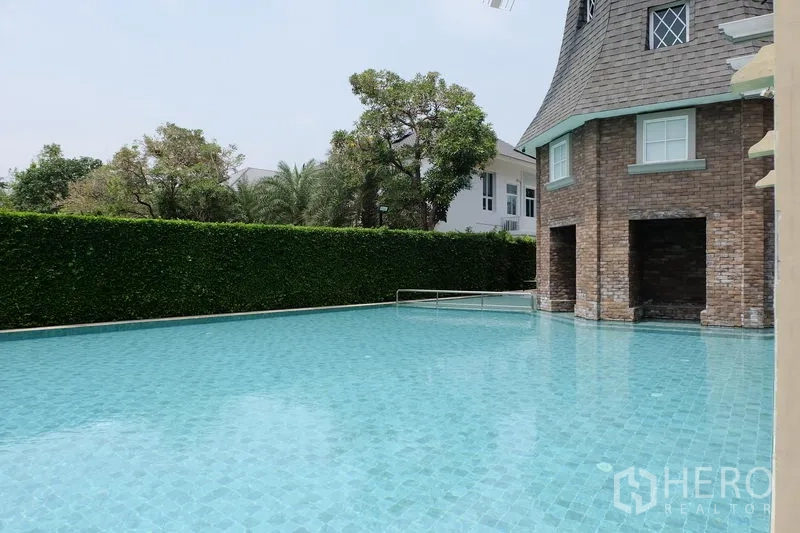 บ้าน ขายในMin Buri, Bangkok - สระว่ายน้ำส่วนกลางขนาดใหญ่ข้างคลับเฮ้าส์ผนังก่ออิฐและแนวรั้วต้นไม้ตัดแต่ง