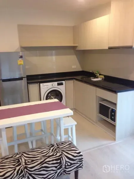 Condominium for rent in Sathon, Bangkok - Compact L-shaped kitchen with black countertops, fridge, washer, microwave and a dining nook.