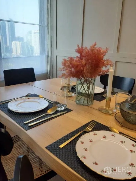 คอนโด ให้เช่าในRatchathewi, Bangkok - มุมรับประทานอาหารสว่างไสว จัดโต๊ะพร้อมภาชนะ ช้อนส้อมทอง แจกันดอกไม้ และวิวเมืองผ่านผ้าม่านโปร่ง