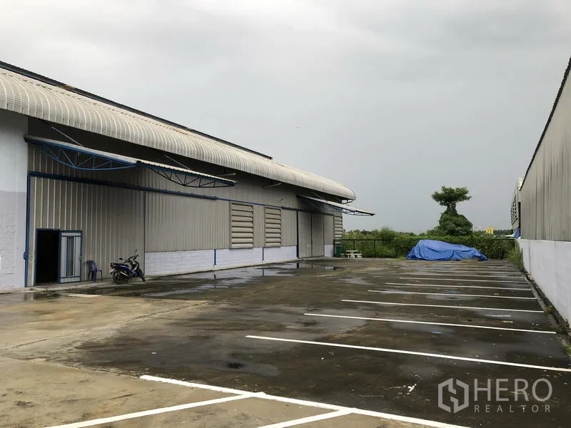Factory for sale in Pathum Thani - Large concrete yard with marked parking bays beside a metal‑clad warehouse building.