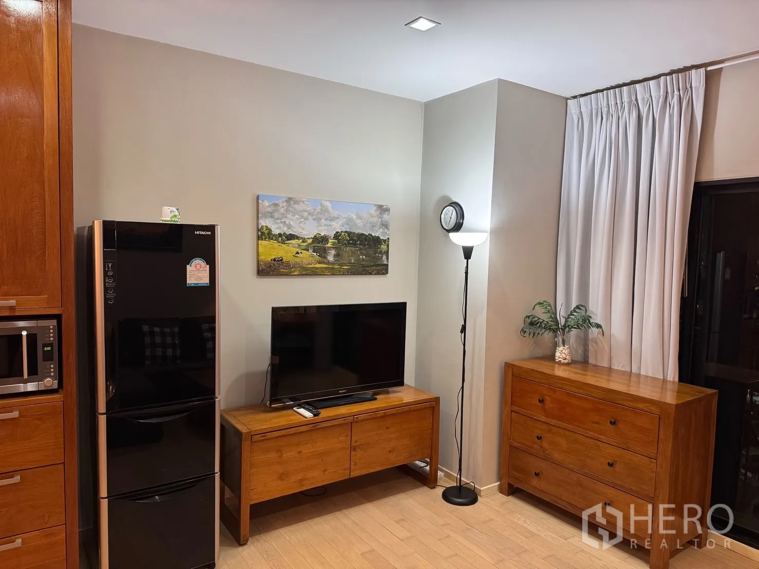 คอนโด for rent or sale in Watthana, Bangkok - ผนังทีวีพร้อมตู้วางทีวีไม้ ตู้เย็นสีดำ ลิ้นชักไม้ และโคมไฟตั้งพื้นข้างผ้าม่านสีอ่อน