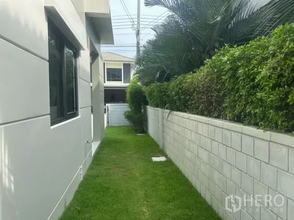 Detached House for sale in Samut Prakan - Side yard with trimmed hedge and boundary wall beside the home’s windows.