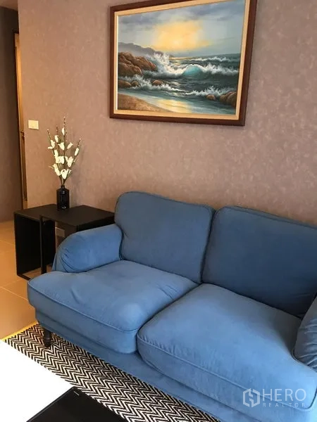 Condominium for rent in Watthana, Bangkok - Cozy seating corner with blue fabric sofa, framed seascape painting and side table over a patterned rug.
