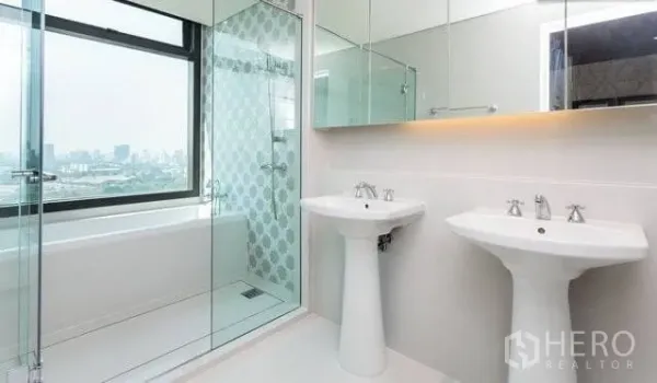Condominium for rent or sale in Khlong Toei, Bangkok - Modern bathroom with twin pedestal sinks, glass shower and bathtub, city view.