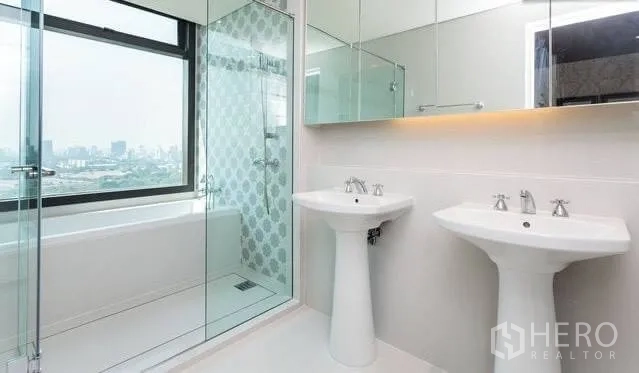 Condominium for rent or sale in Khlong Toei, Bangkok - Modern bathroom with twin pedestal sinks, glass shower and bathtub, city view.