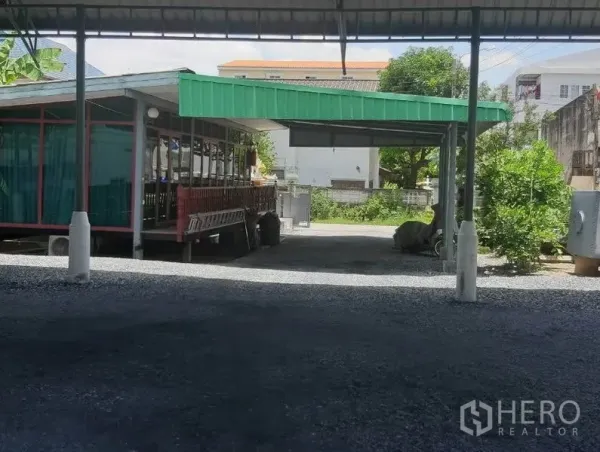 Land for sale in Mueang Samut Prakan, Samut Prakan - Open gravel yard on the plot with a metal canopy and green-roof pavilion, trees and neighboring buildings in view.
