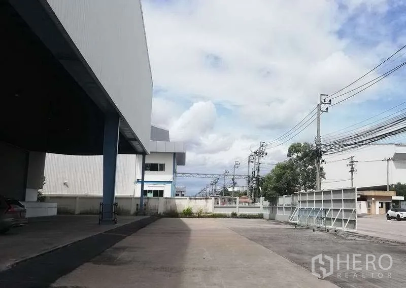 Warehouse for rent in Bang Phli, Samut Prakan - Open driveway and yard beside the warehouse with boundary walls, power lines, and gated access.