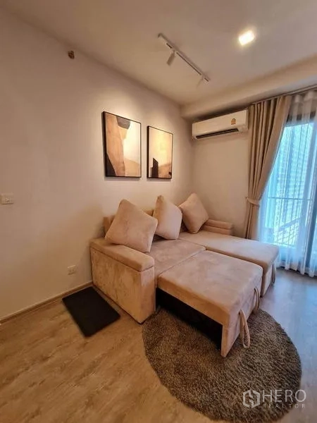 คอนโด ให้เช่า - มุมนั่งเล่นอบอุ่น โซฟา L-Shape พรม ภาพงานศิลป์ แอร์ และประตูกระจกออกสู่ระเบียง