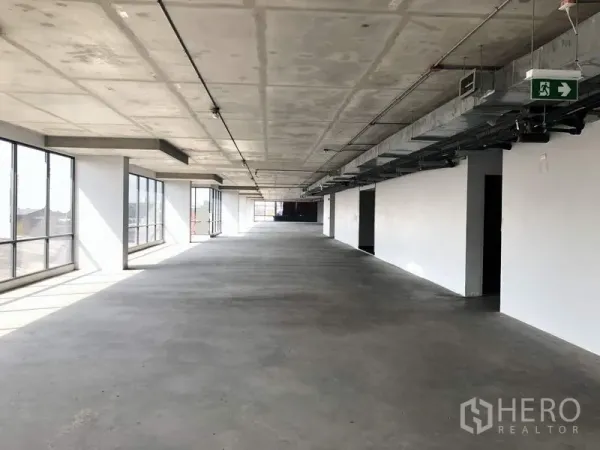 Office Space for rent in Suan Luang, Bangkok - Long open office floor with concrete slab ceiling, emergency exit sign, and continuous windows along one side.