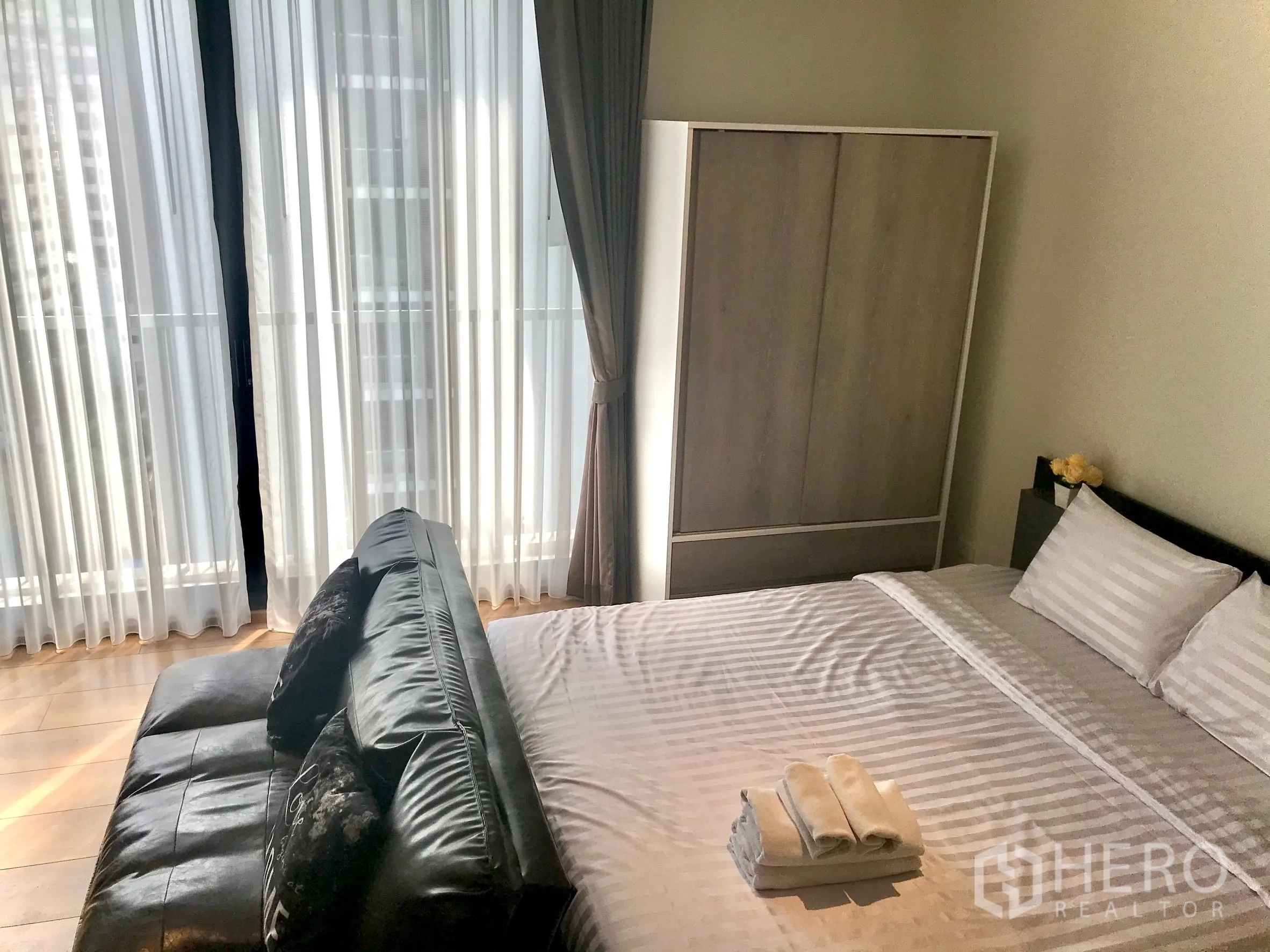 Condominium for sale in Khlong Toei, Bangkok - Bright bedroom with floor-to-ceiling windows, black leather sofa, and a sliding-door wardrobe.