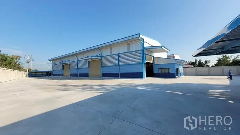 工厂 出租或出售在Phanat Nikhom, Chon Buri - 蓝白配色的现代厂房外观，宽阔混凝土场地与围墙。
