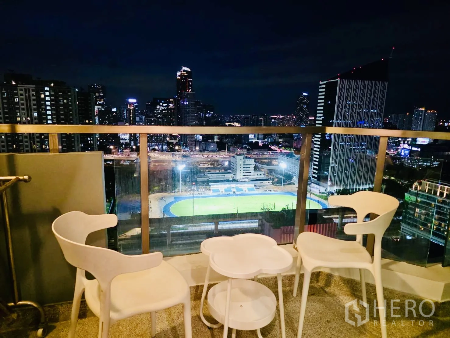Condominium for rent in Huai Khwang, Bangkok - Night view from balcony with chairs facing illuminated city skyline and stadium.
