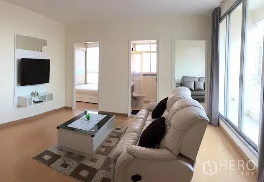 คอนโด ให้เช่าในWatthana, Bangkok - ห้องนั่งเล่นสว่าง โซฟาหนัง ทีวีติดผนัง โต๊ะกลาง และหน้าต่างบานใหญ่เชื่อมสู่สองห้องนอน