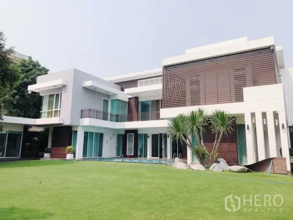 บ้าน ขายในMin Buri, Bangkok - ด้านหน้าอาคารโมเดิร์น สนามหญ้าตัดแต่งและต้นปาล์ม พร้อมหน้าต่างบานใหญ่และระเบียง