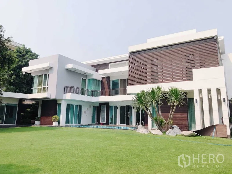 บ้าน ขายในMin Buri, Bangkok - ด้านหน้าอาคารโมเดิร์น สนามหญ้าตัดแต่งและต้นปาล์ม พร้อมหน้าต่างบานใหญ่และระเบียง