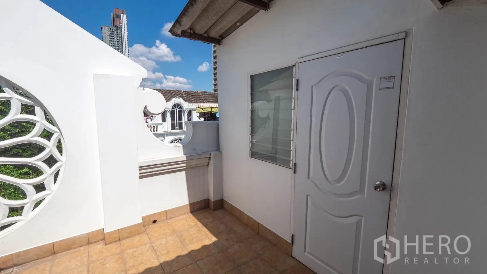Townhouse for rent in Watthana, Bangkok - Upper-floor terrace with tiled floor, decorative circular lattice wall, and white door under a blue sky.