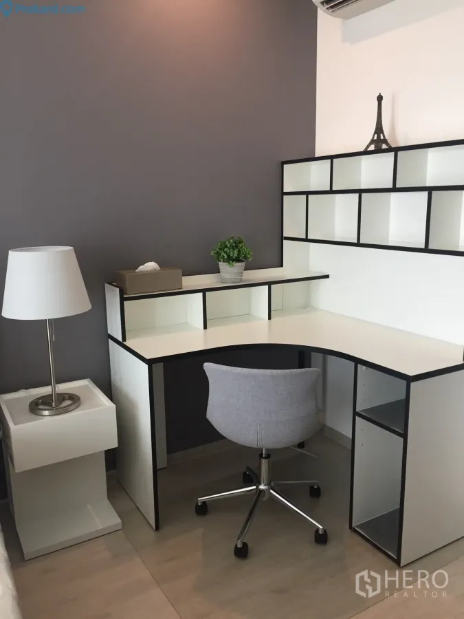 Condominium for rent in Ratchathewi, Bangkok - Compact workspace with L-shaped desk, open shelving, grey swivel chair and lamp against a two-tone wall.
