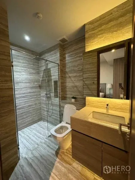 Condominium for rent in Watthana, Bangkok - Modern bathroom with stone tiles, glass-enclosed shower, illuminated vanity, and toilet.