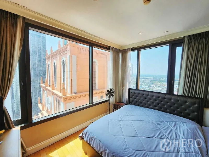 Condominium for sale in Khlong Toei, Bangkok - Corner bedroom with tufted headboard and wraparound windows facing city and ornate facade.