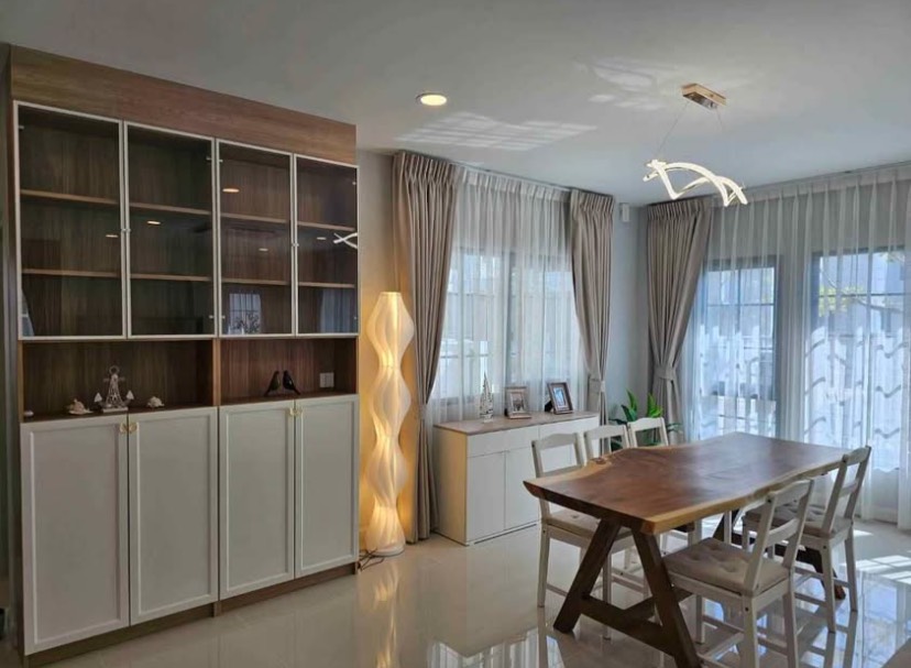 Detached House for rent in Bang Phli, Samut Prakan - Dining space with wooden table, white chairs, display cabinet, pendant light and floor lamp by wide windows.