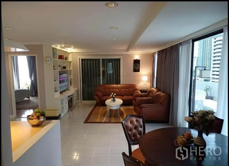 คอนโด ให้เช่าหรือขายในKhlong Toei, Bangkok - ห้องนั่งเล่นคอนโดบนสุขุมวิทสว่างโปร่ง โซฟาหนังสีน้ำตาล ชั้นวางบิวท์อิน และประตูกระจกสไลด์สู่ระเบียงวิวเมือง