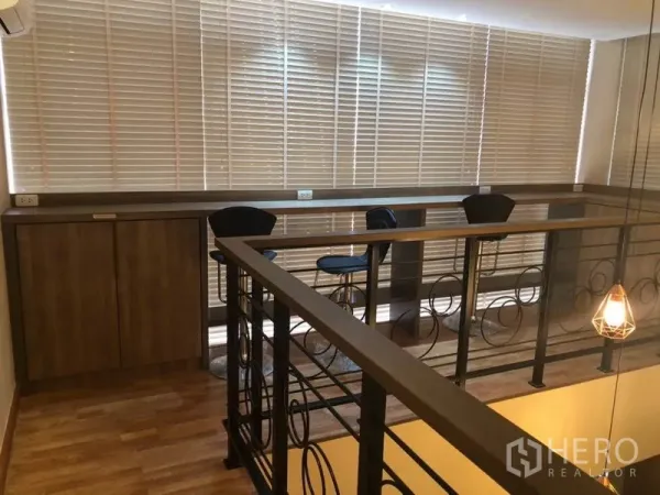 Condominium for sale in Bang Na, Bangkok - Mezzanine workspace with bar counter, stools, wood cabinetry and metal railing beside tall windows.