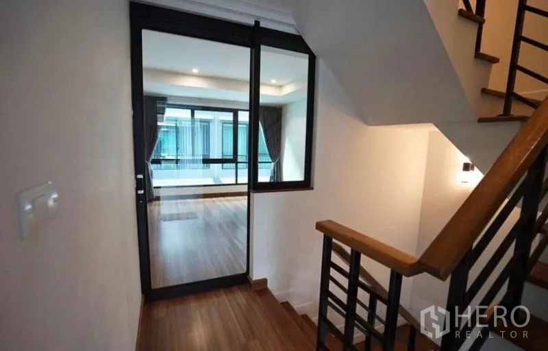 Townhouse for rent in Bang Kapi, Bangkok - Stair landing with wood flooring leading to a bright room through a black-framed glass door.