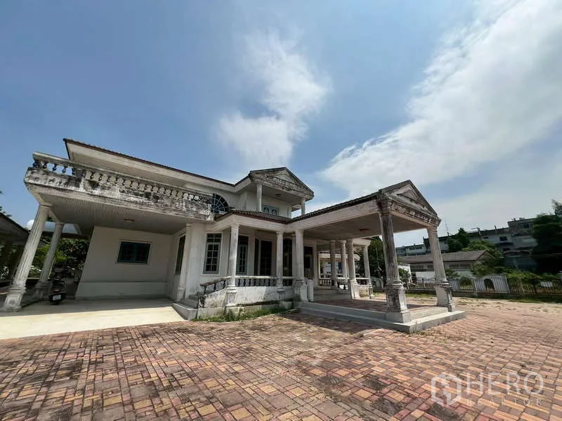 Detached House for rent in Prawet, Bangkok - Two‑storey house with classical columns, tiled driveway and carport under blue sky.