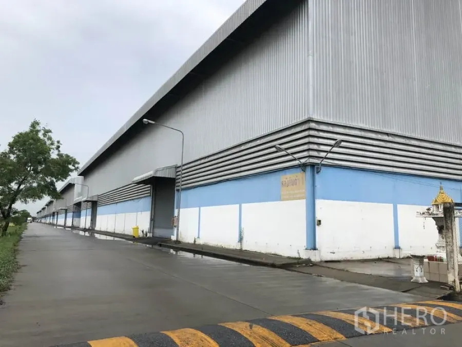 货栈 出租在Phra Nakhon Si Ayutthaya - 仓库侧立面，可见多扇卷闸门与沿混凝土道路的路灯。