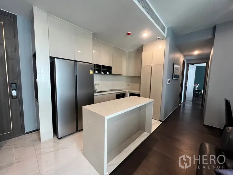 Condominium for rent in Watthana, Bangkok - Open-plan kitchen with white island, glossy cabinetry, double-door fridge and corridor leading to bedrooms.