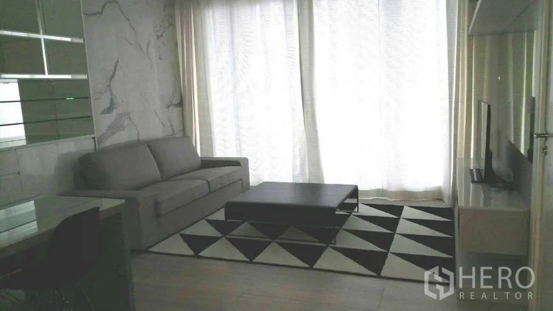 Condominium for rent in Pathum Wan, Bangkok - Cozy living area with TV unit, geometric rug, and sheer curtains filtering natural light.